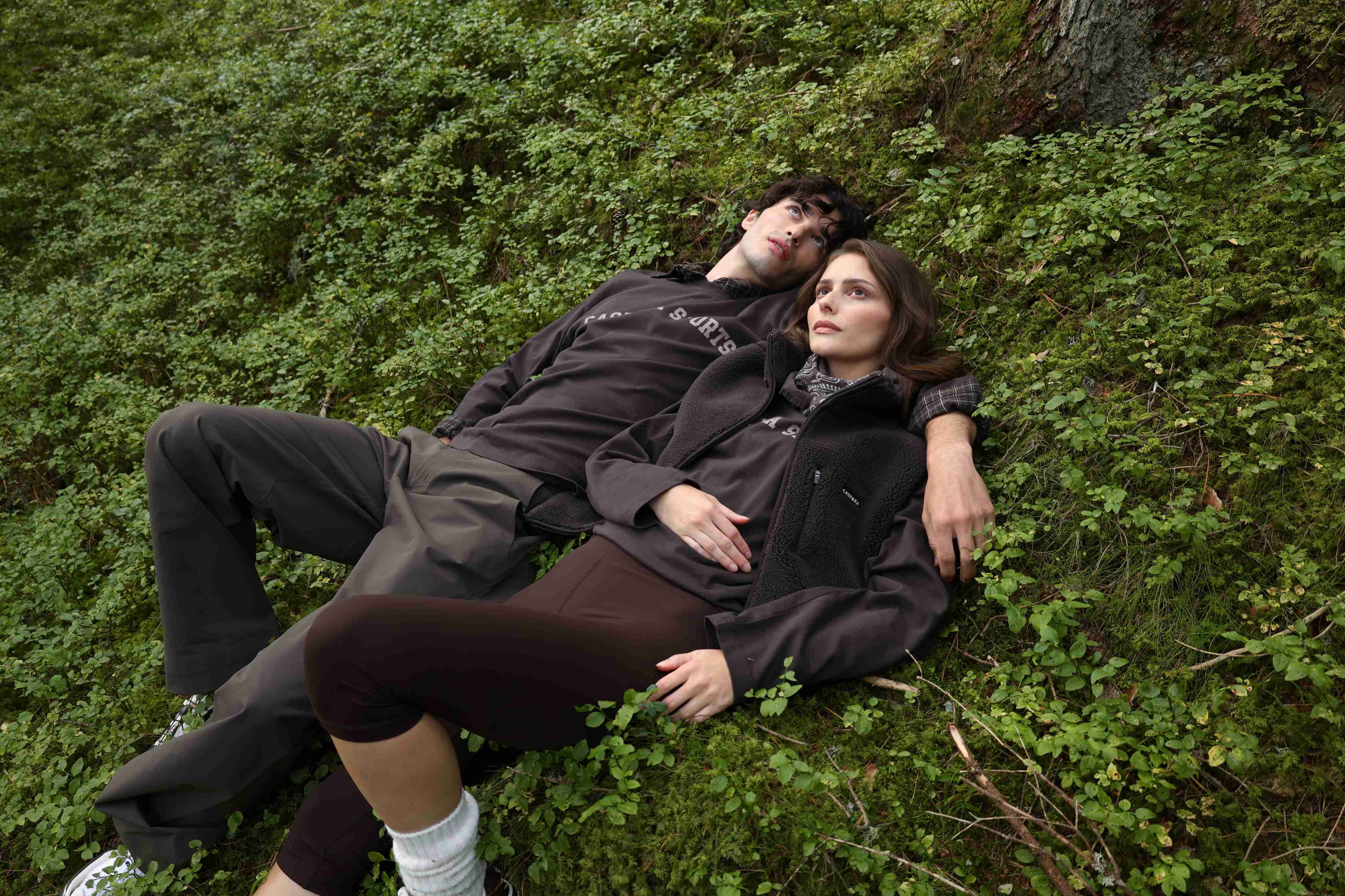 Man and woman wearing espresso CASPARA fleece vest and long sleeve shirts lying on mossy ground.